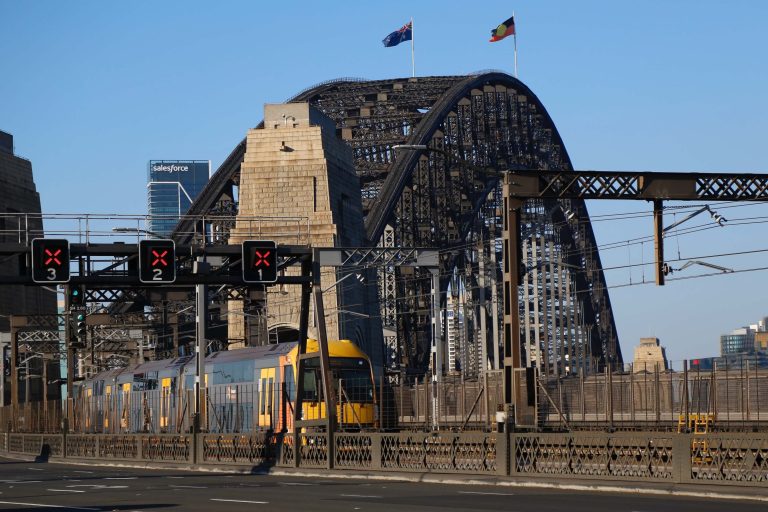 A train travels across the Sydney Harbour Bridge, viewed from the pedestrian footpath on the northern side of the bridge